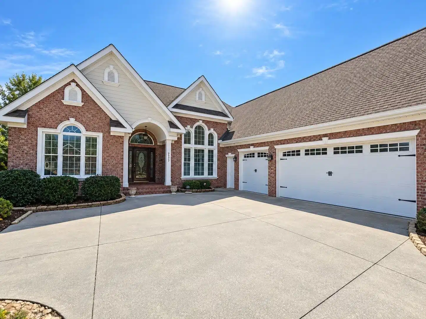 Exterior House Painting Red brick residential home with white-painted trim, columns, and garage doors on concrete driveway.